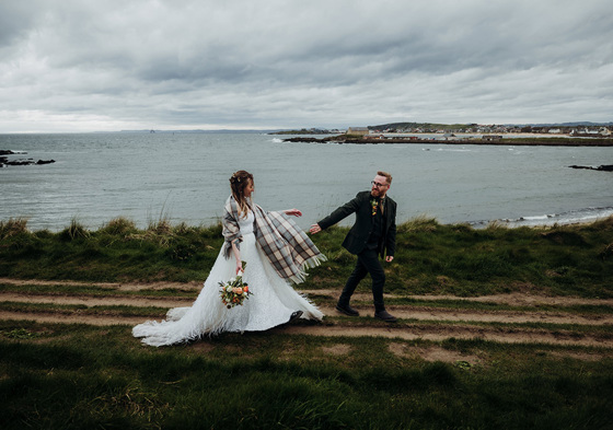 Bride with tartan shawl holds her groom's hand beside the sea with bouquet in other hand